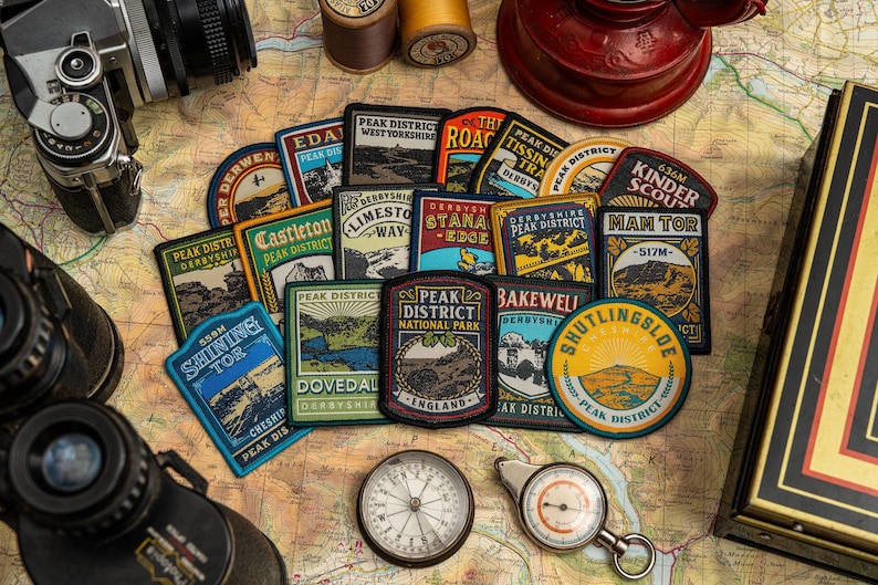 K&ouml;nnte beinhalten: Eine Sammlung farbenfroher Peak District-Patches mit verschiedenen Designs und Texten wie "Kinder Scout" und "Shining Tor". Die Patches sind auf einer Karte zusammen mit einem Kompass, einem Fernglas und einer Vintage-Kamera angeordnet.