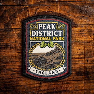Op de afbeelding: Geborduurde patch met de tekst "PEAK DISTRICT NATIONAL PARK ENGLAND". De patch heeft een rechthoekige vorm met een zwarte rand, een blauwe achtergrond en een gedetailleerde illustratie van een landschap met een berg en een krans.
