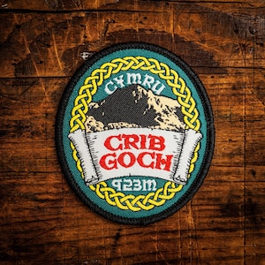 May include: An embroidered patch with a mountain scene and the text "CYMRU" and "CRIB GOCH". The patch is oval-shaped with a black border, a green background, and yellow Celtic knot detailing. The text "923m" is also included.