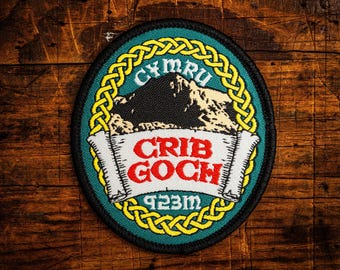 Może przedstawiać: Okrągła, haftowana naszywka z górskim krajobrazem i napisami "CYMRU" i "CRIB GOCH". Naszywka ma czarną obwódkę, zielone tło i żółty celtycki wzór węzła. Zawiera również tekst "923m".