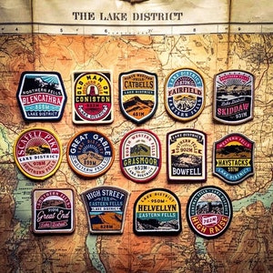 May include: A collection of 14 colourful round patches with mountain scenes and text. Each patch features a different mountain peak in the Lake District, England, with its name and elevation in metres. The patches are arranged in a grid pattern on a map.