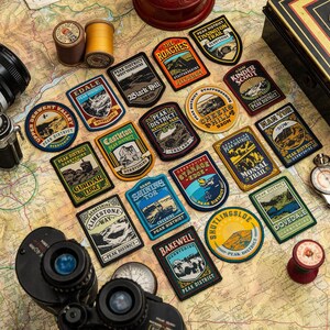 K&ouml;nnte beinhalten: Eine Sammlung von Peak District-Patches, eine Vintage-Kamera, ein Fernglas und ein Kompass sind auf einer Karte angeordnet. Die Patches zeigen verschiedene Orte mit Texten wie "Edale" und "Kinder Scout". Weitere Artikel sind Garnrollen und eine kleine Metallbox.