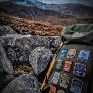 May include: A green backpack with a yellow strap and several patches attached. The patches are colorful and feature names of mountains and their elevations in feet. The backpack is sitting on a pile of rocks in front of a mountain range.
