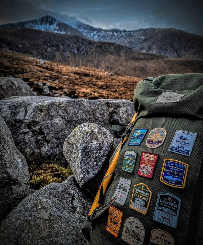 May include: A green backpack with a yellow strap and several patches attached. The patches are colorful and feature names of mountains and their elevations in feet. The backpack is sitting on a pile of rocks in front of a mountain range.