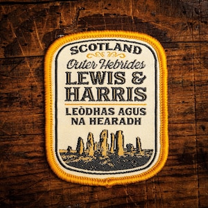 Może przedstawiać: Prostokątna naszywka z żółtą obwódką. Na naszywce widnieje napis "SCOTLAND Outer Hebrides LEWIS & HARRIS" w kolorze czarno-białym. Poniżej widnieje napis "LEÒDHAS AGUS NA HEARADH". Na dole naszywki znajduje się grafika kamieni.