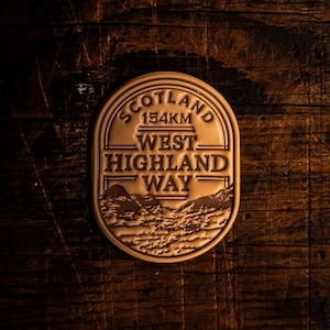 Pode incluir: Um distintivo de metal de cor bronze com o texto "SCOTLAND 154KM WEST HIGHLAND WAY" em forma oval retangular. O distintivo apresenta uma cordilheira na parte inferior. O distintivo está sobre um fundo de grão de madeira escura.