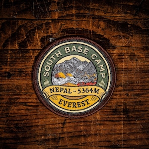 May include: A circular embroidered patch featuring a mountain landscape with the text SOUTH BASE CAMP, NEPAL - 5364M, and EVEREST. The design includes a green border, a yellow banner, and a dark brown edge, showcasing detailed stitching throughout.