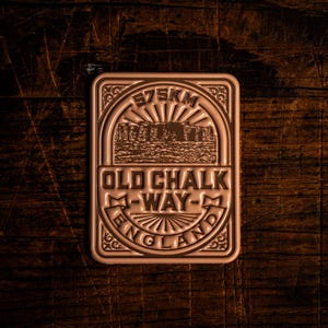 Pode incluir: Placa retangular de cor bronze com o texto "575KM" no topo. A placa apresenta uma ilustração de um monumento e as palavras "OLD CHALK WAY ENGLAND". A placa está sobre um fundo de grão de madeira escura.