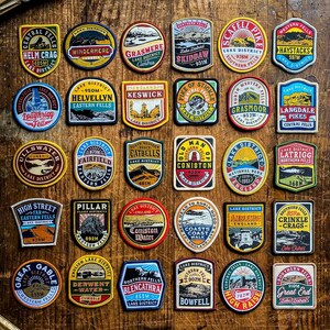 May include: A collection of colorful embroidered patches featuring various landmarks and locations in the Lake District National Park in England. Each patch includes the name of the location, its elevation in meters, and the words "Lake District".