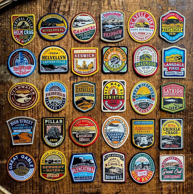 May include: A collection of colorful embroidered patches featuring various landmarks and locations in the Lake District National Park in England. Each patch includes the name of the location, its elevation in meters, and the words "Lake District".
