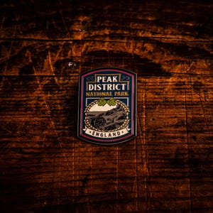 Op de afbeelding: Een rechthoekige emaille pin met de tekst "PEAK DISTRICT NATIONAL PARK ENGLAND". De pin heeft een donkerblauwe achtergrond met een landschapsillustratie en een decoratieve rand in roze, wit en goud.
