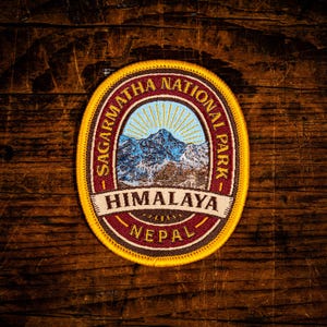 Könnte beinhalten: Ein ovaler gestickter Aufnäher mit einer Berglandschaft unter einer aufgehenden Sonne. Der Text lautet SAGARMATHA NATIONAL PARK, HIMALAYA und NEPAL. Der Aufnäher hat einen leuchtend gelben Rand, einen dunkelroten Innenring und einen hellblauen Himmel.