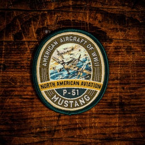 May include: A circular embroidered patch featuring a P-51 Mustang aircraft in flight. The text reads AMERICAN AIRCRAFT OF WWII, NORTH AMERICAN AVIATION, P-51, and MUSTANG. The patch has a dark green border with gold, white, and blue details.