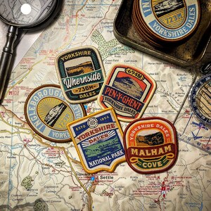 May include: A selection of Yorkshire Dales patches, a compass, and a magnifying glass are displayed on a detailed map. The patches feature mountain illustrations and text like "Whernside" and "Malham Cove". A small metal tray holds additional patches.