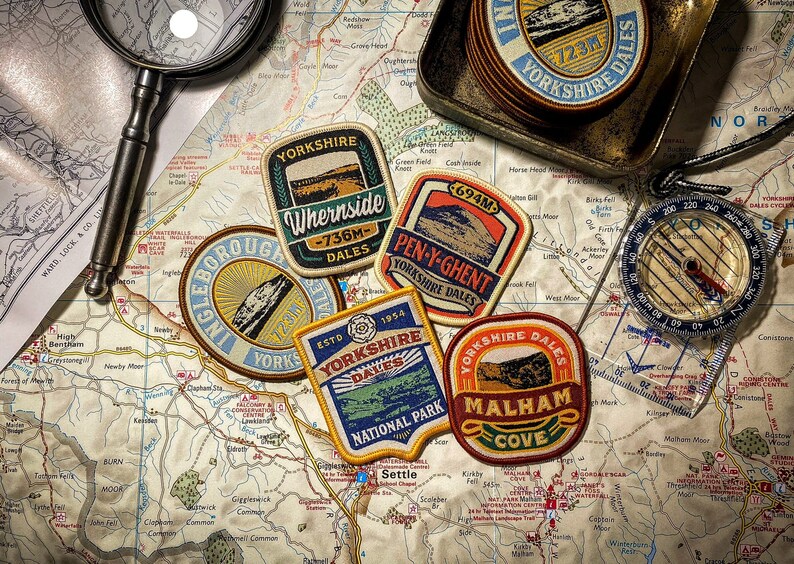 May include: A selection of Yorkshire Dales patches, a compass, and a magnifying glass are displayed on a detailed map. The patches feature mountain illustrations and text like "Whernside" and "Malham Cove". A small metal tray holds additional patches.