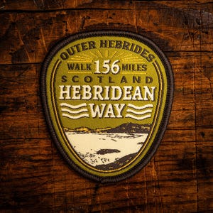 Puede incluir: Parche bordado con borde verde y negro, con el texto "Outer Hebrides Scotland Hebridean Way Walk 156 Miles". El parche representa un paisaje con una masa de agua y montañas. El parche está sobre una superficie de madera.