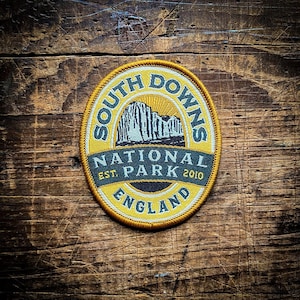 May include: A yellow and white embroidered patch with the text "South Downs National Park Est. 2010 England" and a graphic of a cliffside with a sunrise.