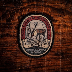 Pode incluir: Patch oval com uma ilustração de rena, texto "Flora & Fauna of the British Isles" e "Eurasian Tundra Reindeer". O patch tem uma borda vermelha, branca e preta. Em um fundo de madeira.