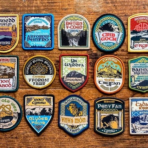 May include: A collection of 16 colourful embroidered patches featuring Welsh landmarks and locations. Each patch has a different design and includes the name of the location, the height of the mountain, or the name of the park. The patches are all made of felt and have a stitched border.