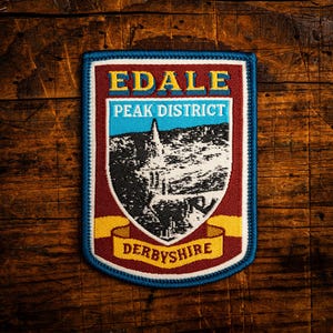 May include: A rectangular patch with a blue border, featuring the text "EDALE PEAK DISTRICT DERBYSHIRE". The patch has a maroon background with a landscape illustration in the center. The text is in yellow and white.