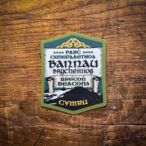May include: A green and yellow sticker with the text "Parc Cenedlaethol Bannau Brycheiniog Brecon Beacons Cymru" and a mountain scene.