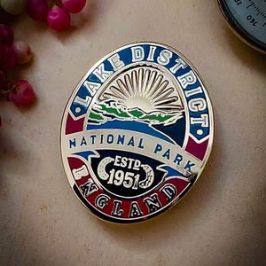 May include: A gold-colored pin with a red, white, and blue design. It says "Lake District National Park England" and "Est. 1951" in a circle.
