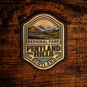 Op de afbeelding: Een patch met de tekst "Regional Park Pentland Hills Scotland". De patch heeft een berg- en waterontwerp met een beige rand. De patch ligt op een houten oppervlak.