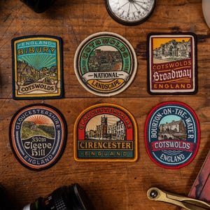K&ouml;nnte beinhalten: Eine Sammlung von bestickten Aufn&auml;hern mit verschiedenen Orten in den Cotswolds, England. Jeder Aufn&auml;her zeigt ein anderes Design, einschlie&szlig;lich Texten wie "Bibury", "Broadway" und "Cirencester" sowie Illustrationen.