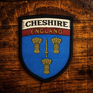 Op de afbeelding: Een patch met de tekst "CHESHIRE ENGLAND". Het blauwe schildontwerp toont drie korenaren en een gouden zwaard. De rand is zwart. De achtergrond is een houten oppervlak.