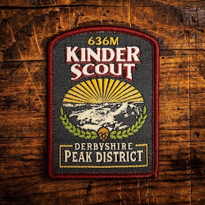 May include: A rectangular patch with the text "636M KINDER SCOUT" in white and red lettering. Below is a yellow sunburst design over a mountain scene, framed by green laurel leaves. The bottom reads "DERBYSHIRE PEAK DISTRICT" in a yellow rectangle.