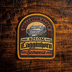 Op de afbeelding: Geborduurde patch met een berglandschap met de tekst "Walliser Alpen" en "4010M Lagginnhorn Schweiz". De patch heeft een zwarte rand met oranje, bruine en gele accenten, tegen een houten achtergrond.