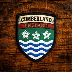 May include: A shield-shaped patch with the words "CUMBERLAND ENGLAND" at the top. The patch features a red and green stripe, three white flowers, and blue and white wavy lines. The patch is bordered in black.