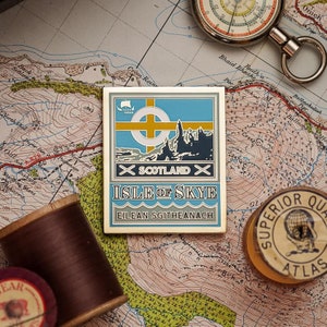 May include: A gold-coloured enamel pin with a blue, white and yellow design. The pin features a stylised image of the Isle of Skye, Scotland, with the text "Isle of Skye" and "Eilean Sgitheanach" below the image. The pin is on a map.