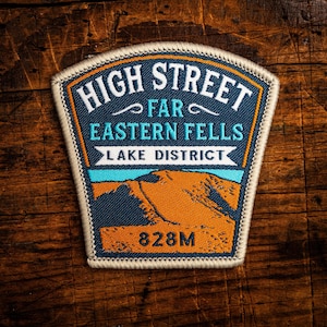 May include: A rectangular patch with the text "HIGH STREET FAR EASTERN FELLS LAKE DISTRICT" in white and blue. The patch features an orange mountain scene and the number "828M". The patch has a white border and is set against a wooden background.