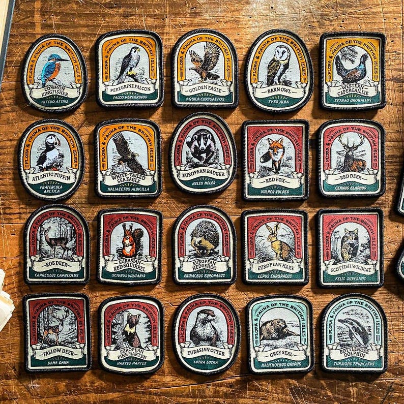 May include: A collection of 20 colourful embroidered patches featuring various animals and plants found in the British Isles. Each patch has a white background with a red, orange, or blue border. The patches are arranged in a grid pattern on a wooden surface.