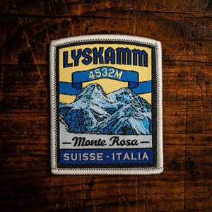 May include: Embroidered patch with the text "LYSKAMM 4532M" and a mountain illustration. The patch also includes "Monte Rosa" and "SUISSE - ITALIA" on a rectangular shape with a black border. The background is a wooden surface.