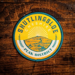 May include: A circular patch with the text "SHUTLINGSLOE CHESHIRE PEAK DISTRICT" in teal and yellow. The patch features a mountain landscape design with a rising sun and a teal border. The patch is on a wooden surface.