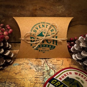 May include: A brown paper package tied with twine and a green logo that reads "The Adventure Patch Company - Est. MMXVII".