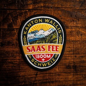 Puede incluir: Parche bordado ovalado con el texto "Kanton Wallis" y "Saas Fee 1800M Schweiz". El parche presenta un diseño de montaña y sol en rojo, amarillo, azul y verde. El parche tiene un borde negro.