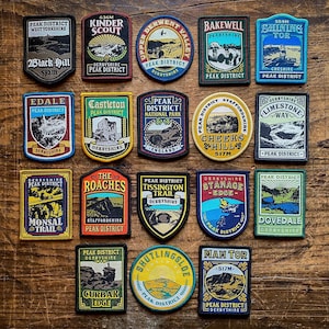 Puede incluir: Una colección de 16 parches bordados coloridos que representan varios puntos de referencia y ubicaciones en el Parque Nacional Peak District en Inglaterra. Los parches son circulares, cuadrados y en forma de escudo y presentan texto como "Kinder Scout", "Shining Tor", "Mam Tor", "Cheeks Hill", "The Roaches", "Monsal Trail", "Curbar Edge", "Dovedale", "Edale", "Castleton", "Tissington Trail", "Stanage Edge", "Bakewell", "Limestone Way" y "Upper Derwent Valley".