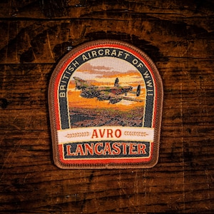 May include: A shield-shaped embroidered patch featuring an Avro Lancaster aircraft flying over a landscape at sunset. The border text reads BRITISH AIRCRAFT OF WWII, with AVRO LANCASTER printed in orange on a white and red background at the bottom.