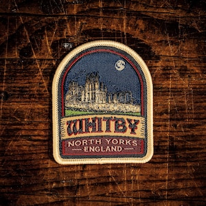 May include: A semi-circular embroidered patch featuring a dark blue night sky with a moon above a stone abbey. The text reads WHITBY in large letters, with NORTH YORKS ENGLAND below on a red background. The patch has a cream-coloured border.