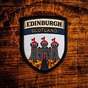 May include: A shield-shaped embroidered patch featuring the text EDINBURGH in black on a cream background and SCOTLAND in gold on a blue band. Below, a white section displays a black stone castle with red roof accents and a rocky base.