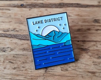 May include: A blue and white enamel pin with the text "LAKE DISTRICT" and a stylized image of a mountain range and a lake with a moon in the sky.