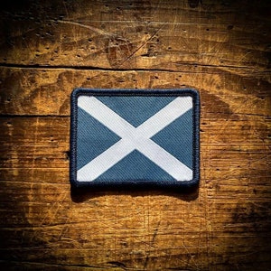 May include: A blue and white Scottish flag patch with a white cross on a dark blue background. The patch is surrounded by a dark blue border and has a stitched edge.