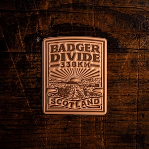 May include: A rectangular, bronze-colored badge with the words "BADGER DIVIDE" and "SCOTLAND" in embossed lettering. The badge also features the text "338KM" and a landscape design with a rising sun. The badge is set against a dark wood grain background.
