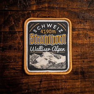 May include: Embroidered patch with a black background, featuring the text "Schweiz 4190m Strahlhorn Walliser Alpen" in yellow and white. The patch has a mountain graphic and a gold border.