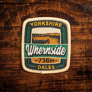 May include: A rectangular patch with the words "Yorkshire" and "Whernside" in white lettering. The patch features a landscape scene and the text "-736M- Dales". The patch has a black border and is set against a wooden background.