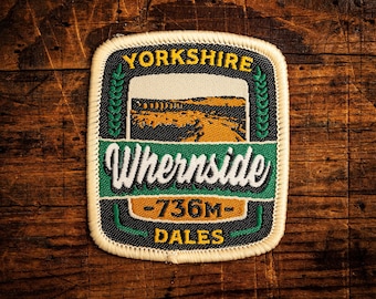 May include: A rectangular patch with the words "Yorkshire" and "Whernside" in white lettering. The patch features a landscape scene and the text "-736M- Dales". The patch has a black border and is set against a wooden background.