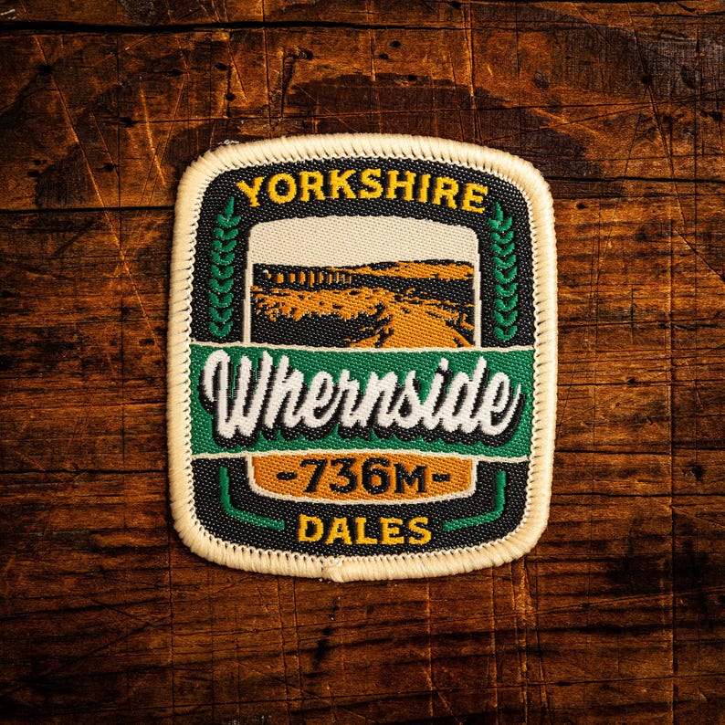 May include: A rectangular patch with the words "Yorkshire" and "Whernside" in white lettering. The patch features a landscape scene and the text "-736M- Dales". The patch has a black border and is set against a wooden background.